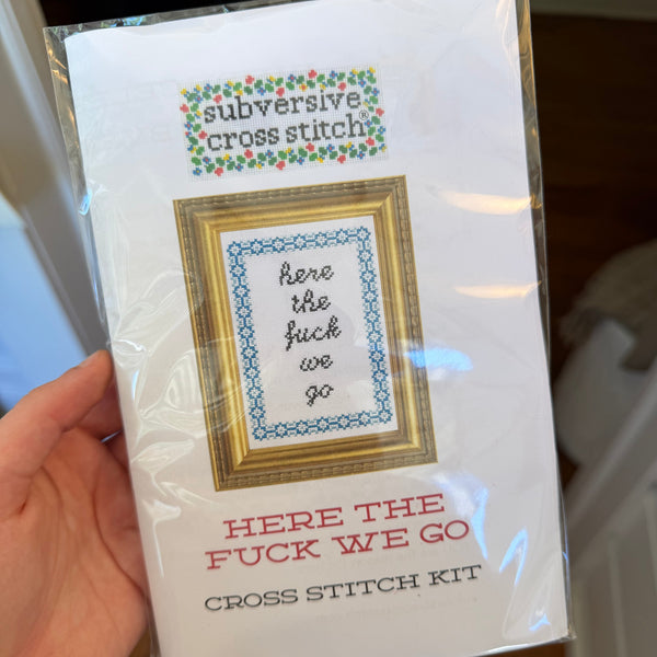 Hand holding a Deluxe Cross Stitch Kit, Here the Fuck We Go, featuring a framed subversive cross stitch pattern with floral accents, perfect for funny home decor or gifts from Knotty and Board, promoting humor and style in any room.