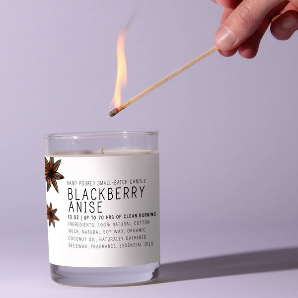 Hand lighting a Just Bee Blackberry Anise, 13 oz candle with a wooden wick in a clear glass jar, perfect for home decor, gifts, and cozy kitchen ambiance from Knotty and Board.