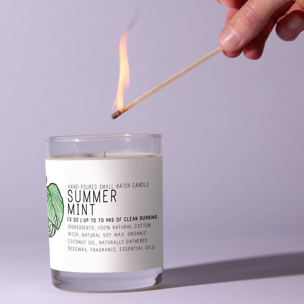 Hand lighting a Just Bee Summer Mint Candle, 13 oz, with a match against a light lavender background, showcasing clean and natural decor for kitchen or dining spaces, perfect gifts from Knotty and Board – Every Room Needs A Little Knotty and Board.