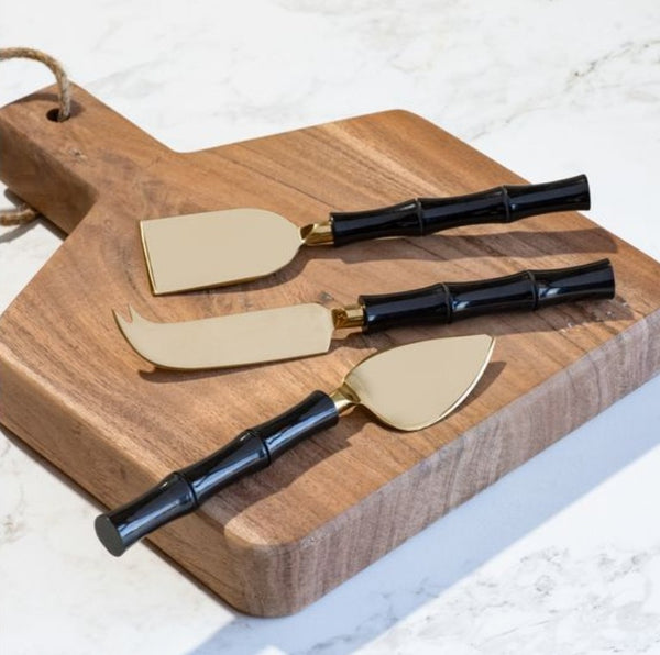 Black Gold Cheese Knife Set with sleek gold blades and black bamboo-style handles displayed on a wooden cutting board, perfect for kitchen decor and entertaining gifts by Knotty and Board.