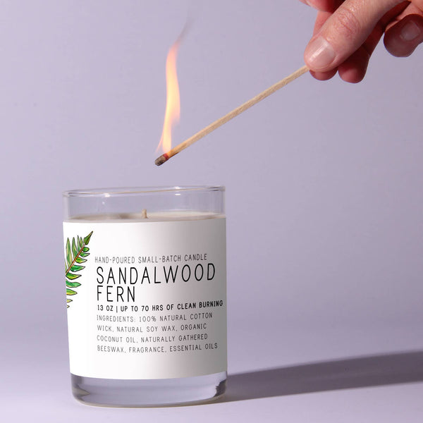 A hand holding a lit match lighting the wick of a Just Bee Sandalwood Fern Candle, 13 oz, in a clear glass jar with a minimalist label featuring green fern decor, ideal for home and kitchen ambiance from Knotty and Board.