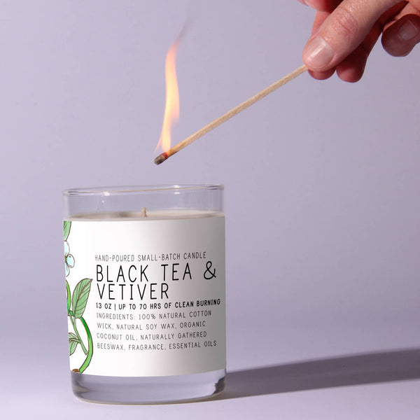 Hand lighting a Just Bee Black Tea Vetiver, 13 oz candle in a clear glass jar with a botanical label, perfect for cozy decor and unique gift ideas from Knotty and Board.
