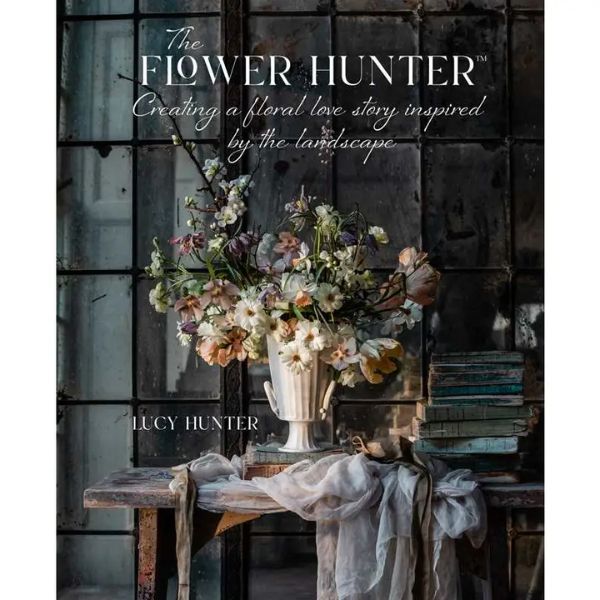 Book cover for "Flower Hunter: Creating A Floral Love Story Inspired By the Landscape" featuring a rustic bouquet of soft pastel flowers in a white vase on a wooden table with draped fabric, stacks of books, and moody window light, reflecting Knotty and Board's curated decor and gift style.