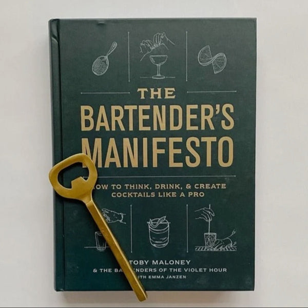 The Bartender's Manifesto book with a dark cover featuring cocktail illustrations, paired with a gold bottle opener, ideal for kitchen decor and entertaining gifts from Knotty and Board.